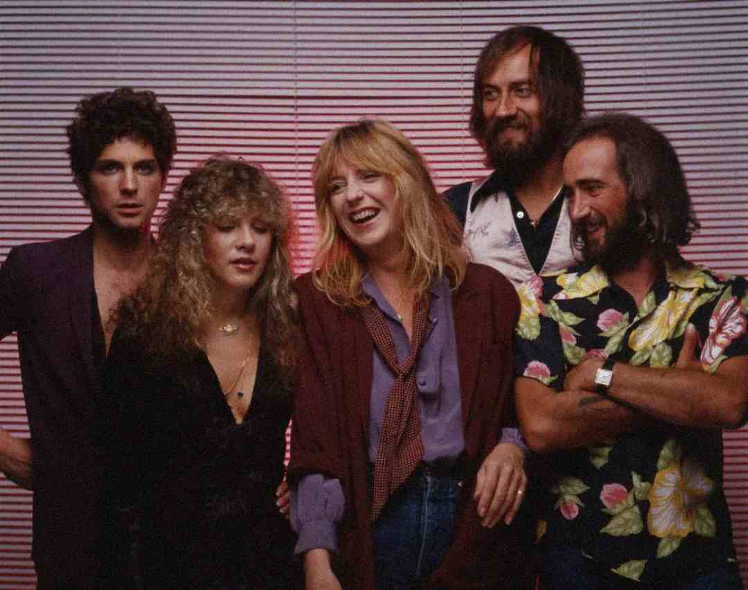 Five people stand together in front of pink-striped blinds, smiling and laughing. Three men and two women are dressed in casual 1970s-style clothing, with relaxed poses and friendly expressions.