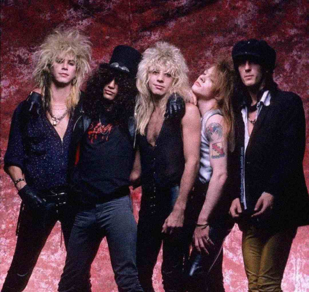 Five members of a rock band with big 1980s-style hair stand together in front of a maroon backdrop, wearing leather, hats, and accessories, striking relaxed, confident poses.