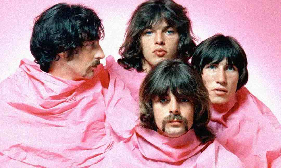 Four men with long hair and mustaches are wrapped in pink fabric, posing closely together against a pink background. Their expressions are serious and contemplative.