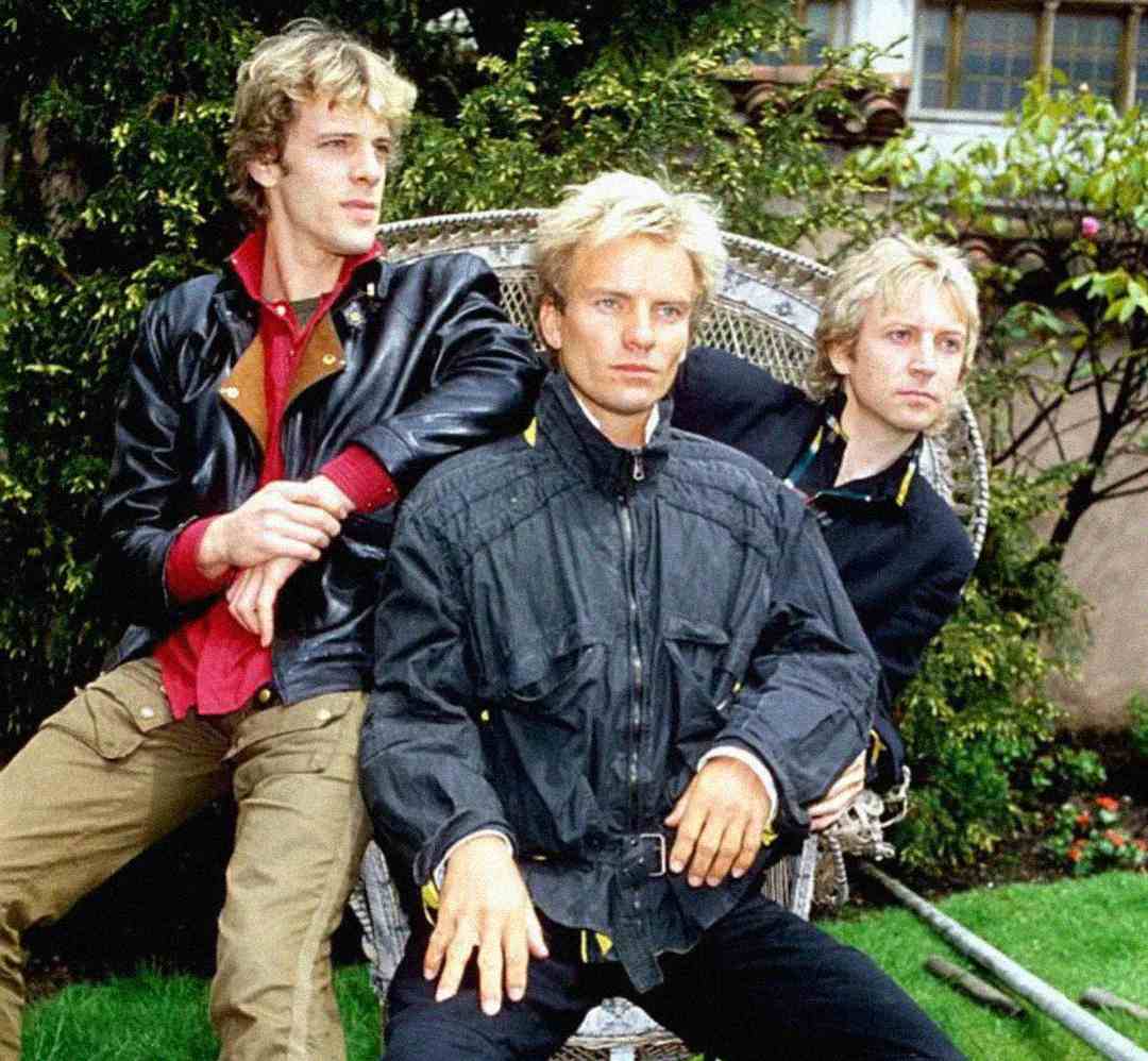 Three men with blond hair sit and pose outdoors; one sits in a wicker chair while the other two stand and sit beside him. They wear casual jackets and look serious. Lush greenery and a building are visible in the background.