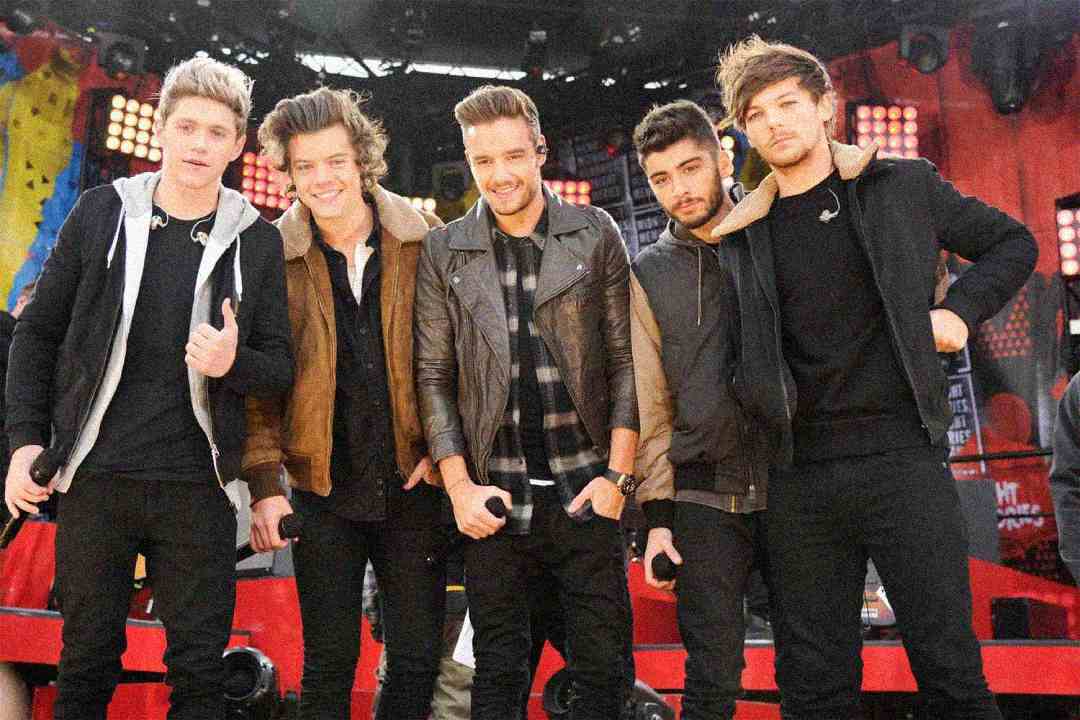 Five young men stand close together on an outdoor stage, smiling and holding microphones, with colorful lights and equipment in the background. They are dressed in casual jackets and jeans.