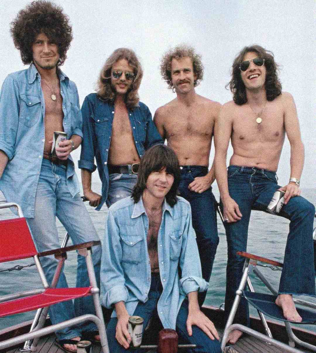 Five men on a boat, some shirtless and some in blue denim shirts and jeans, smiling and holding drinks, with water visible in the background and red and white chairs in the foreground.