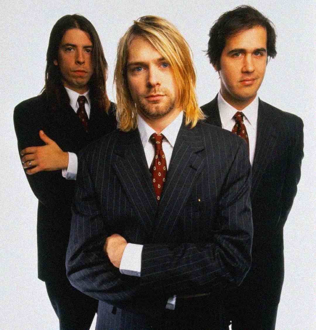 Three men wearing dark pinstripe suits, white shirts, and patterned ties stand in a studio with a plain light background. The man in front has long blonde hair; the two men behind have dark hair and serious expressions.