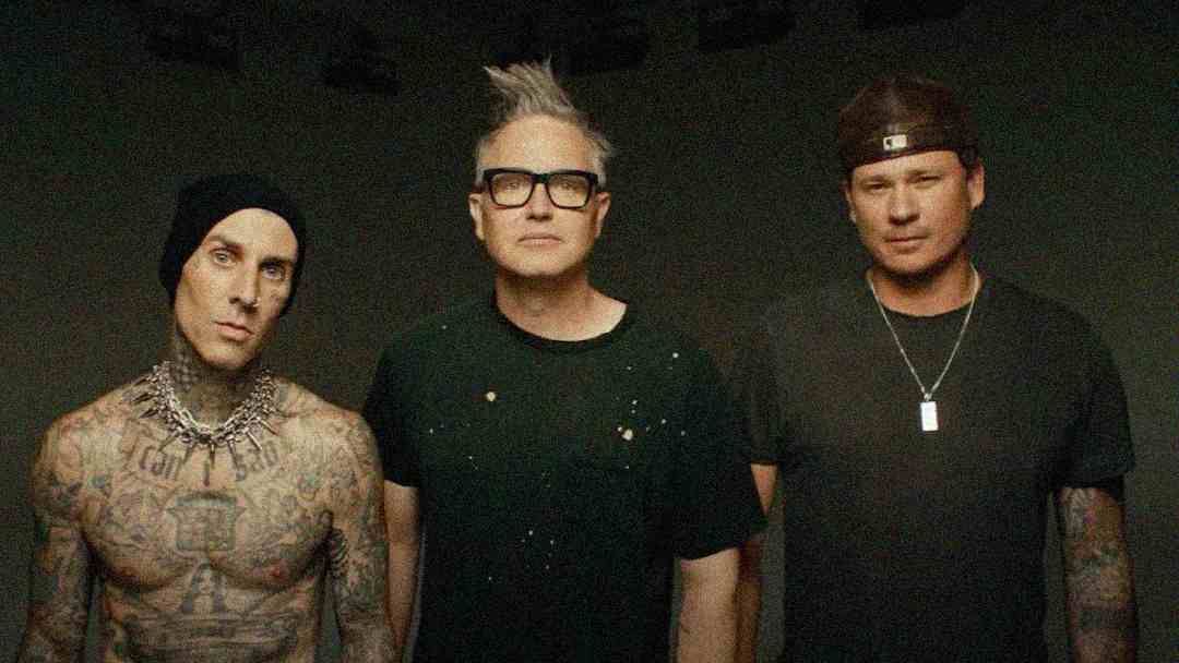 Three men stand side by side against a dark background. The man on the left has visible tattoos, wears a beanie and necklace, the middle man has glasses and gray hair, and the right man wears a backwards cap and a necklace.