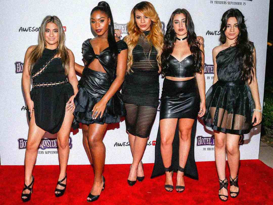 Five women stand together on a red carpet, posing for a photo. They are all wearing stylish black dresses and heels. A movie premiere backdrop is behind them, featuring logos and titles.
