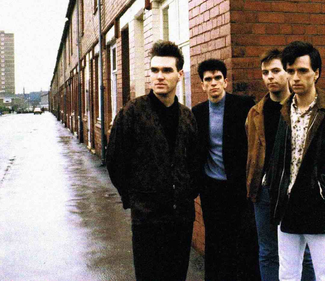 Four men stand on a wet, empty street next to a row of brick buildings, looking serious. The sky is overcast and a tall building is visible in the background.