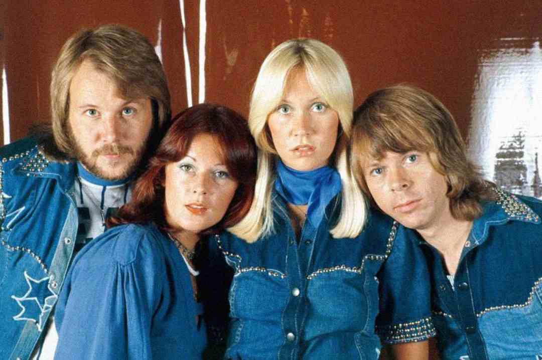 Four people in matching blue outfits stand close together against a reddish-brown background, looking directly at the camera. Two have long blonde hair and two have long reddish or brown hair.