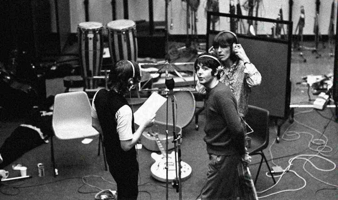 Three musicians wearing headphones stand around microphones in a recording studio, with music stands, chairs, instruments, and cables scattered around them. The scene has a casual, creative atmosphere.