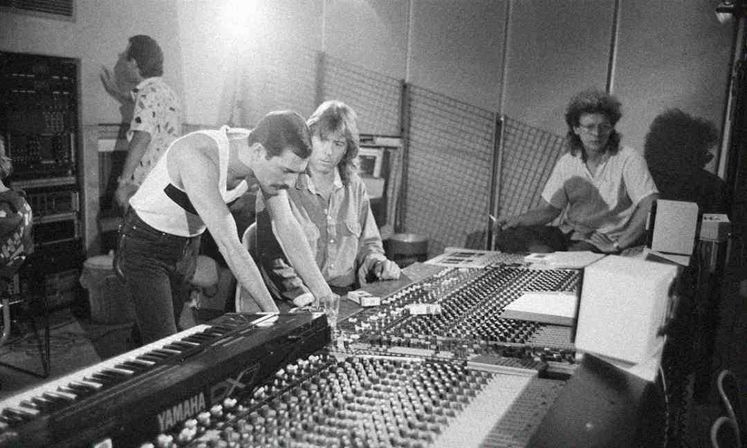 Black and white photo of three people in a music recording studio, with two men focused on a large mixing console and another man sitting behind them. A keyboard is visible in the foreground.