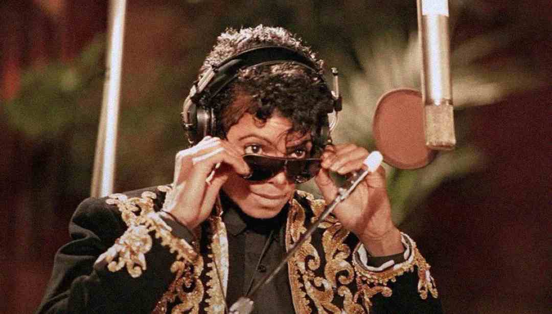 A person in a black and gold embroidered jacket and headphones lifts their sunglasses while standing in front of a large studio microphone. The background is blurred with warm colors and greenery.