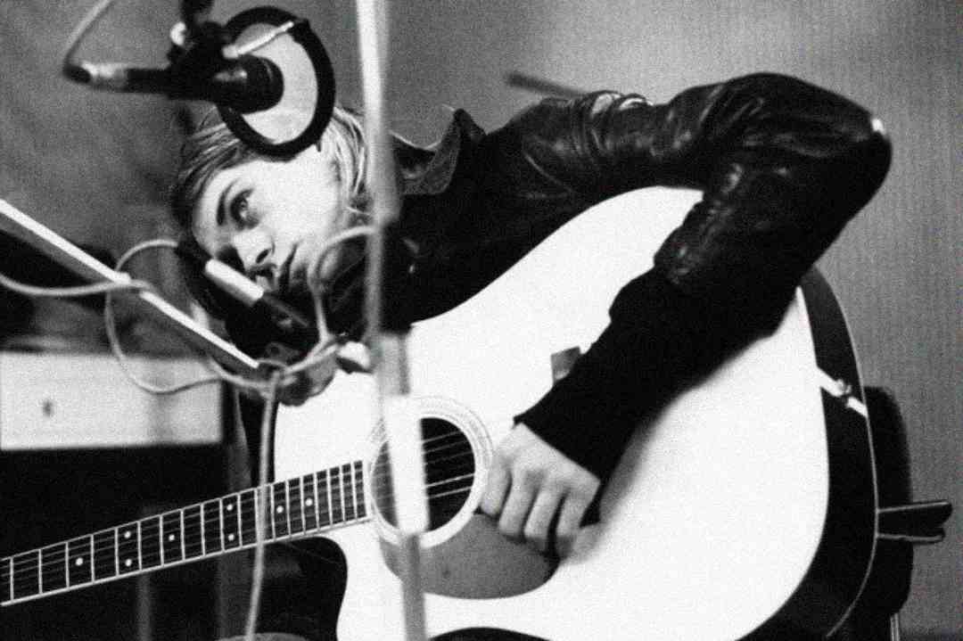 A person with long hair and a leather jacket leans over an acoustic guitar, resting their head near a microphone in a recording studio. The image is in black and white.