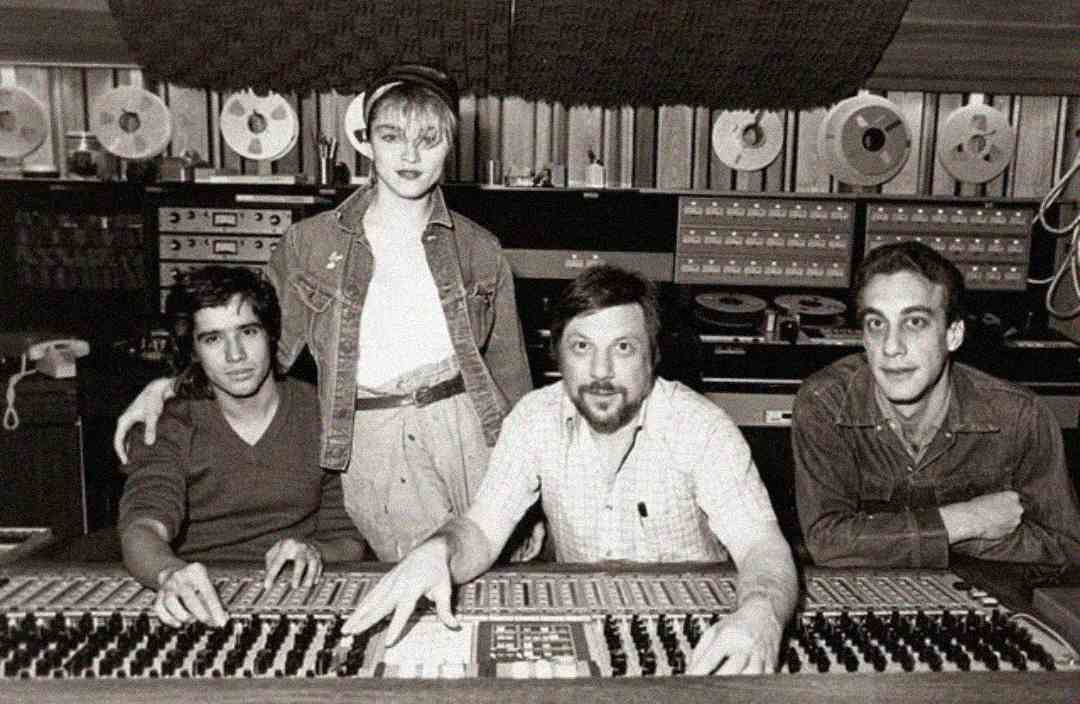 A black-and-white photo shows three men and one woman in a recording studio, with audio equipment and tape reels in the background. The woman stands behind the men, who are seated at a large mixing console.