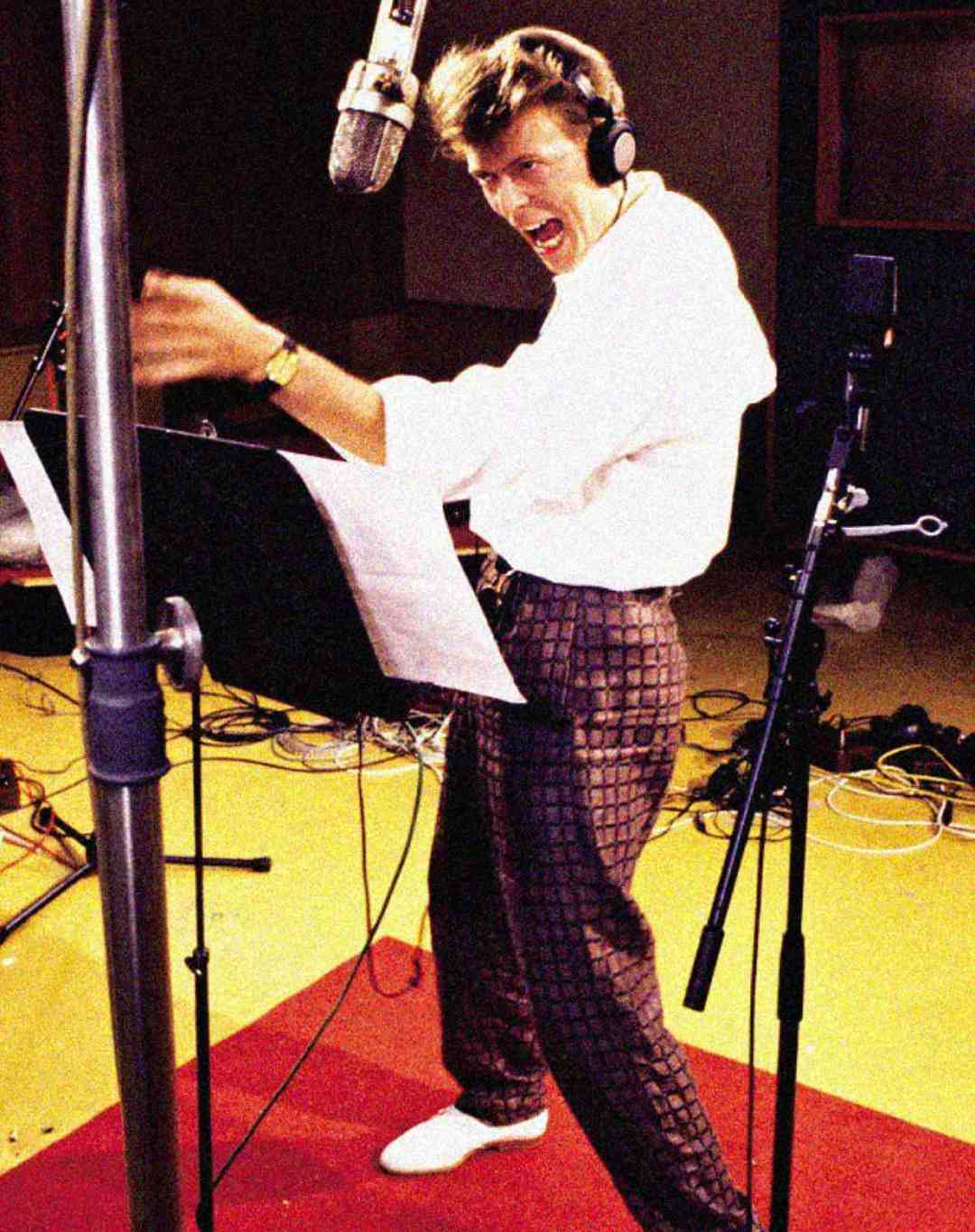 A man wearing headphones and patterned pants passionately sings into a studio microphone, standing by a music stand, with cables and recording equipment visible in the background.
