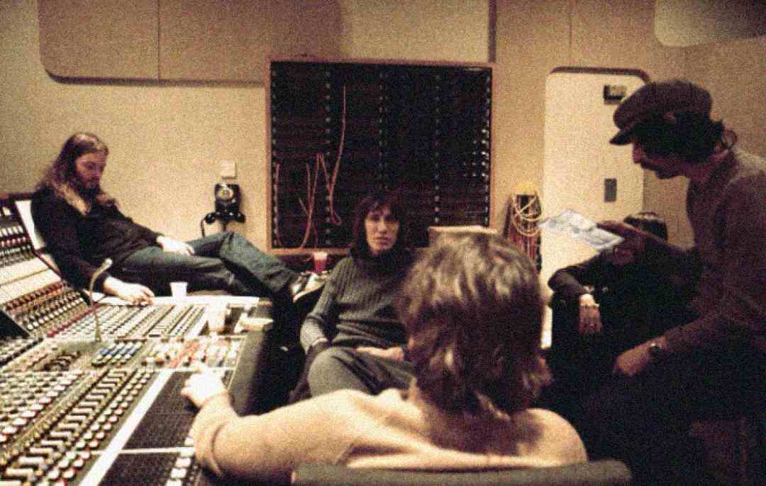 Four people are sitting and talking in a recording studio. Audio equipment and a large mixing console are visible in the foreground, with cables and panels on the wall behind them. The mood appears relaxed and focused.