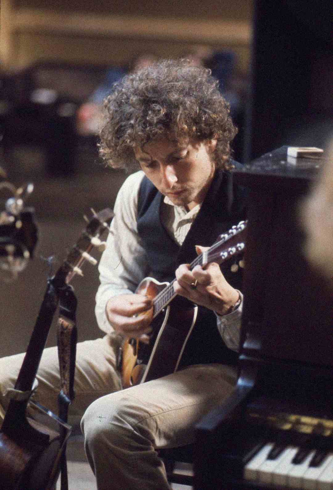 A man with curly hair and a vest sits indoors, playing a mandolin. Several instruments, including a piano and guitars, surround him. The setting appears calm and focused.