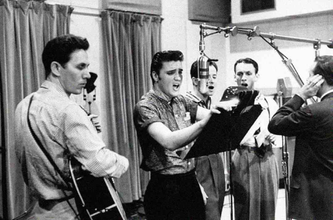 A group of four men in a recording studio, with one man singing passionately into a vintage microphone while others accompany him; one plays guitar, and curtains hang in the background.