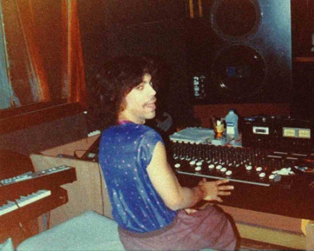 A person in a blue sleeveless shirt sits at a recording studio mixing console, looking back at the camera and smiling. Large speakers, keyboards, and recording equipment surround them.