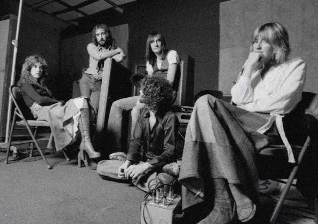 A black-and-white photo of five people in a relaxed indoor setting. Four are seated on chairs, while one sits cross-legged on the floor. They all appear thoughtful, dressed in casual 1970s-style clothing.