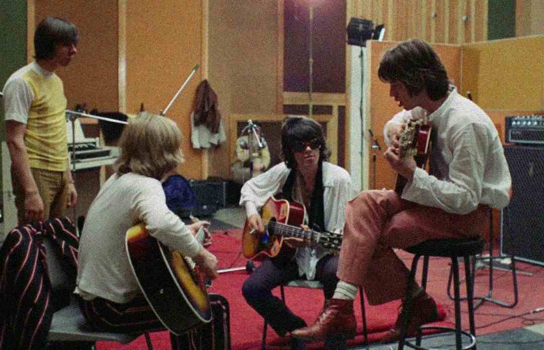 Four men are in a music studio with wood-paneled walls. Three are seated and playing acoustic guitars, while one stands nearby. Cables, speakers, and recording equipment can be seen around them.
