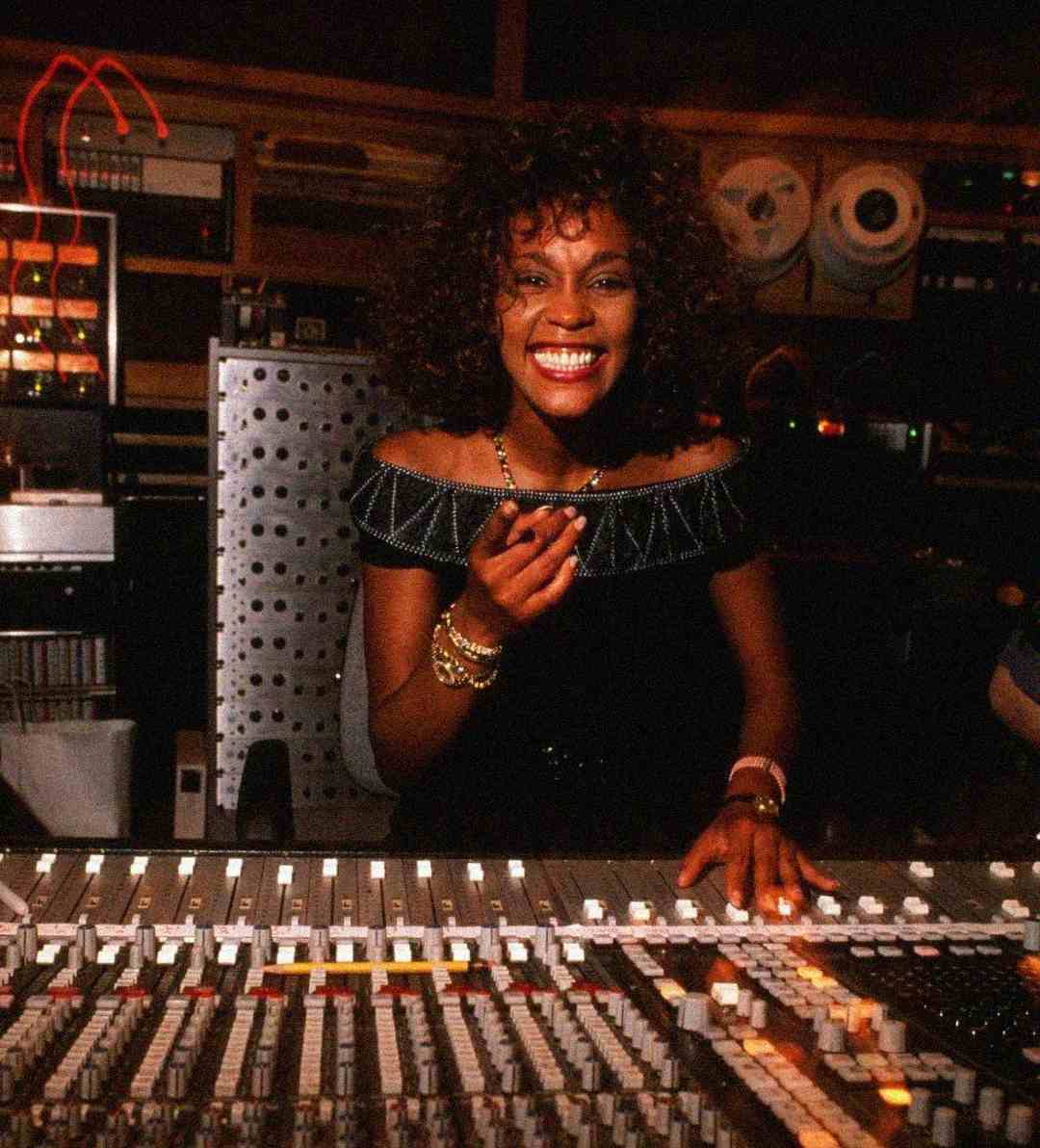 A woman with curly hair and a big smile sits at a mixing console in a recording studio, wearing an off-the-shoulder black dress and jewelry, surrounded by audio equipment and reels.