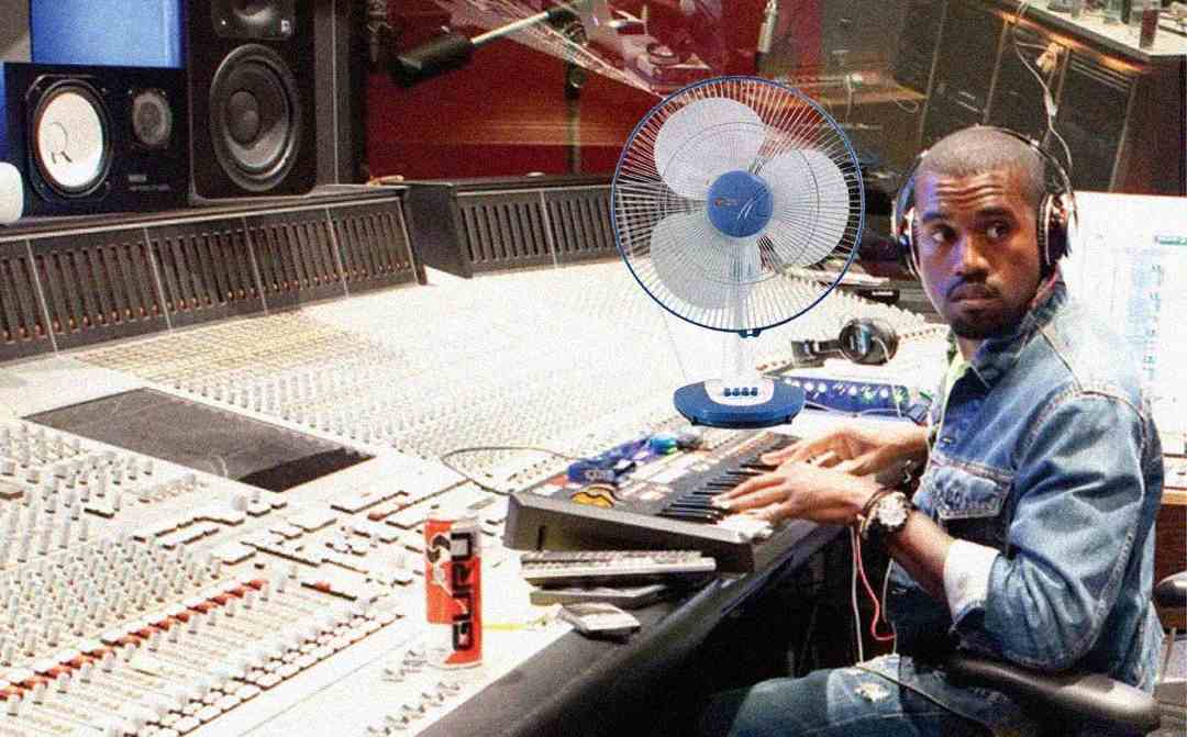 A man wearing headphones sits at a large recording studio mixing console, playing a keyboard. A table fan and an energy drink can are on the desk. The man looks to his right, appearing focused.