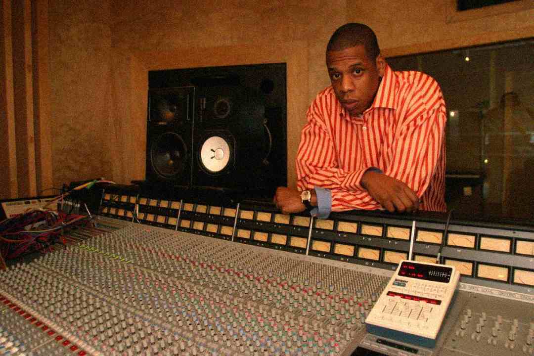 A man in a red and white striped shirt leans on a large audio mixing console in a recording studio, with speakers and sound equipment visible in the background.
