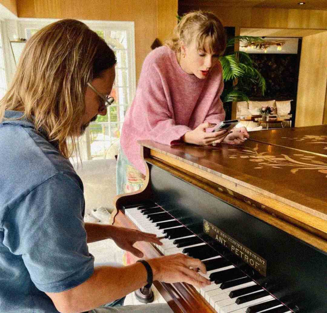 A man with long hair plays a grand piano while a woman in a pink sweater leans on the piano, holding a phone and looking at its screen. They are in a bright room with plants and large windows.