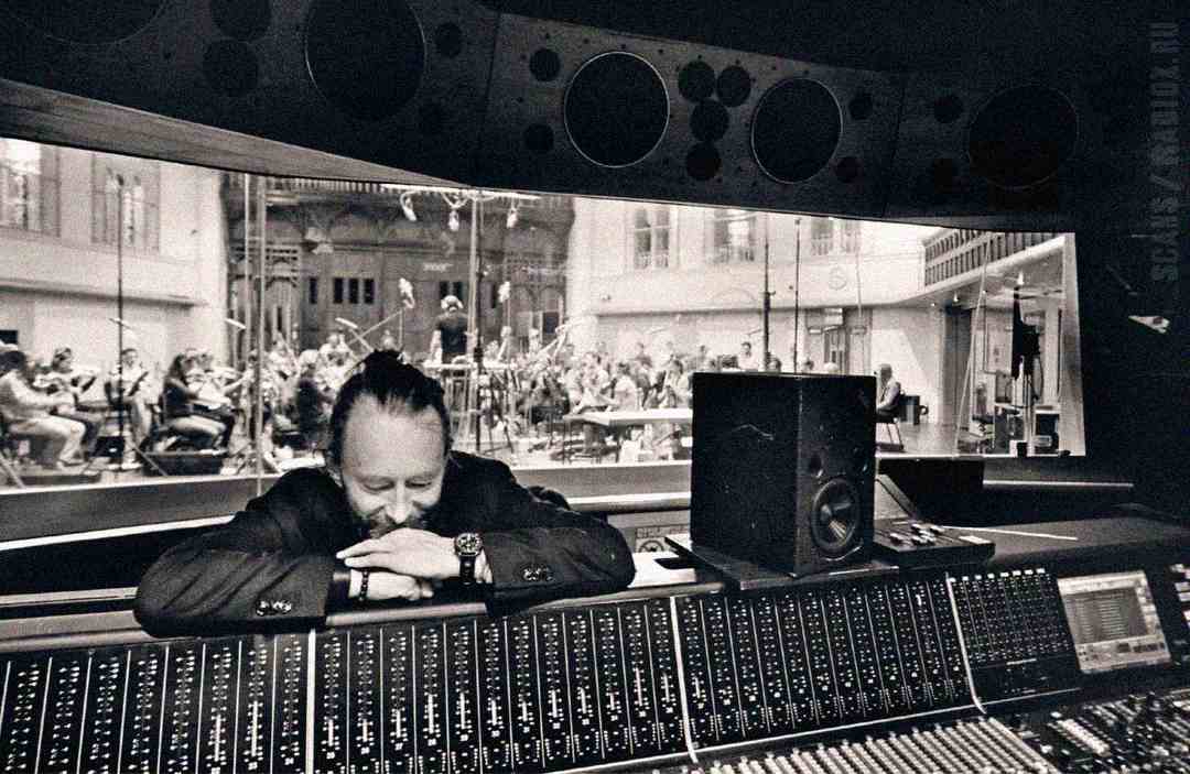 A person with their arms folded and head resting on a recording studio mixing desk, smiling, with an orchestra visible through a large window in the background. The image is in black and white.