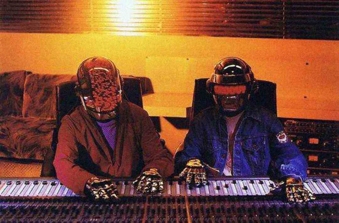 Two people wearing futuristic helmets and gloves sit at an audio mixing console in a dimly lit recording studio, focusing on the sound equipment in front of them.