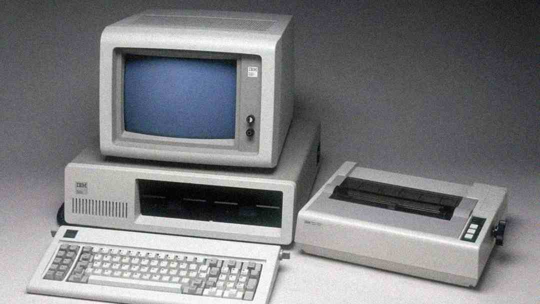 An old IBM personal computer with a CRT monitor, separate keyboard, and a dot matrix printer, all in beige color, displayed on a gray surface.