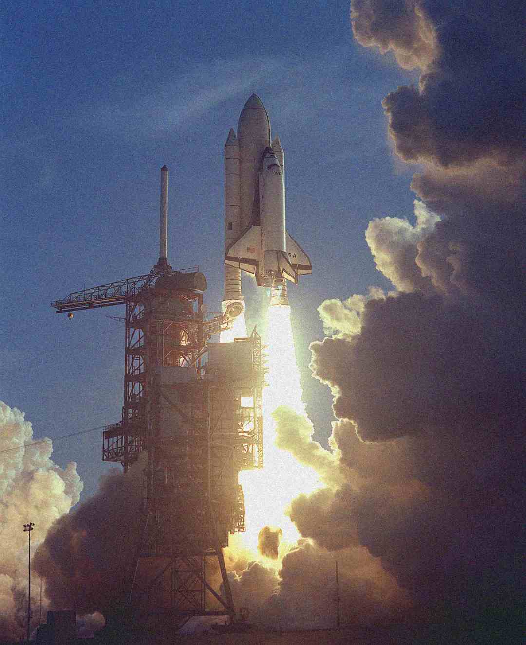 A space shuttle launches from its pad, emitting bright flames and smoke as it ascends into the sky. Large clouds of exhaust billow around the launch structure in daylight.