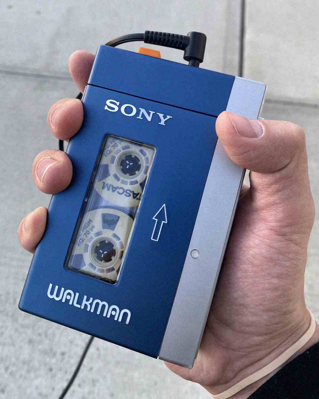 A hand holds a blue and silver Sony Walkman with a visible cassette tape inside, plugged in with headphones. The background shows a gray sidewalk.