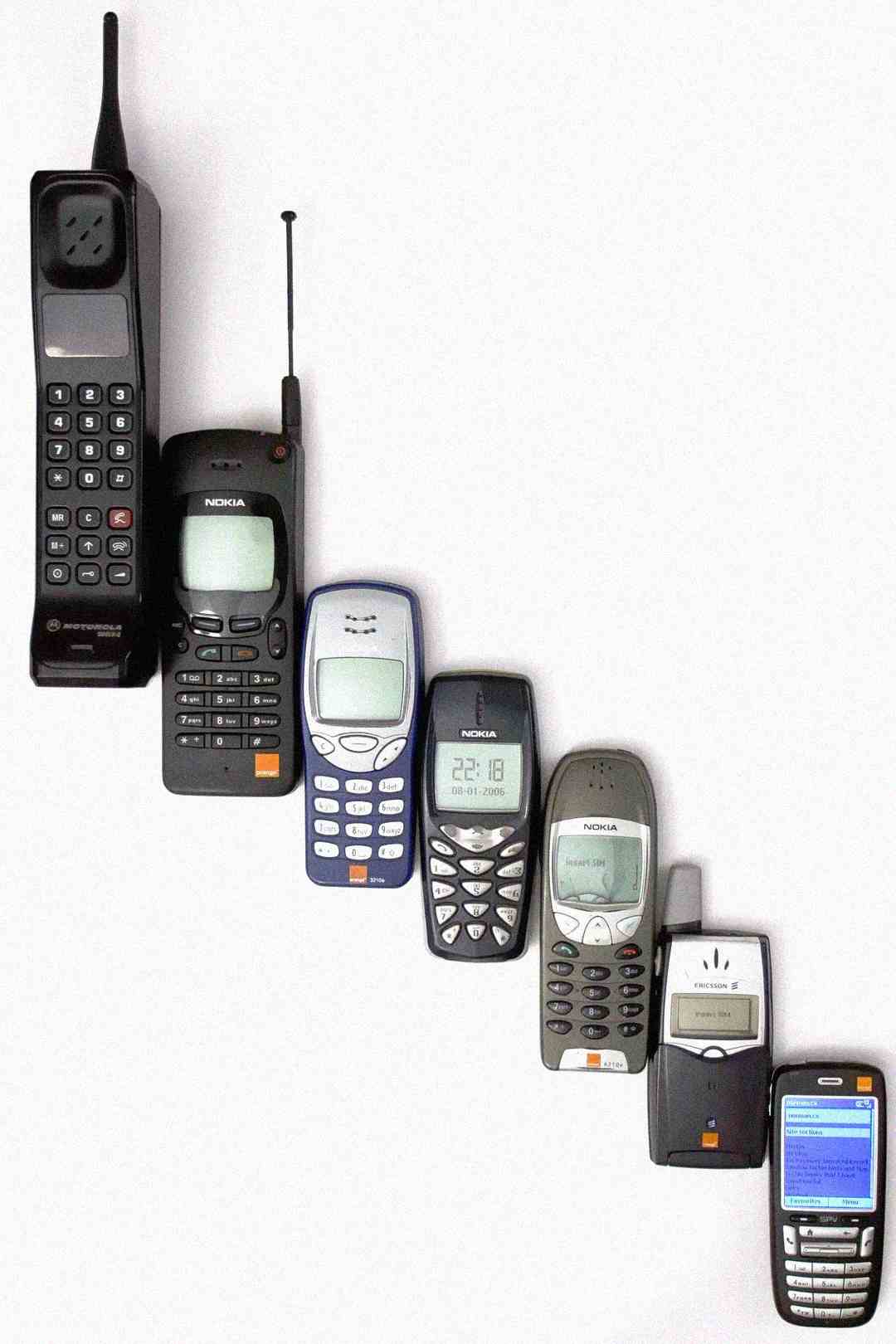Seven mobile phones are arranged in a row from left to right, showing the evolution from a large, early model with an antenna to smaller, more modern models with displays and keypads.