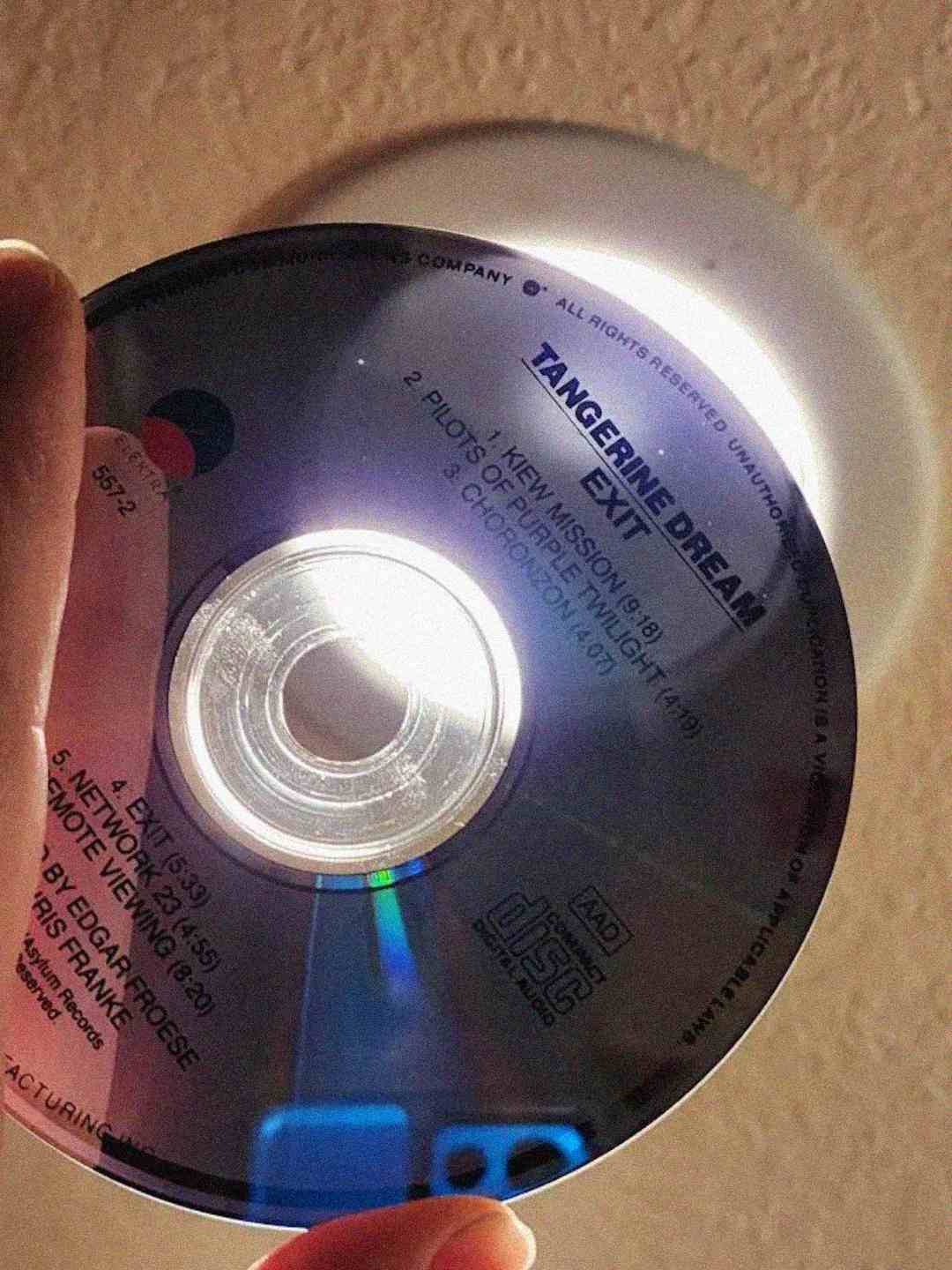 A hand holds a Tangerine Dream "Exit" CD near a light source, causing the disc to reflect light and reveal its tracklist. The background is a beige wall and a round ceiling light.