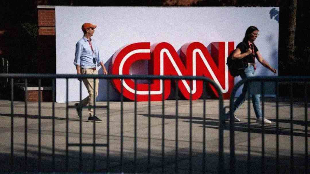 Two people walk in front of a large red CNN logo displayed on a white wall outdoors, with metal barricades in the foreground.