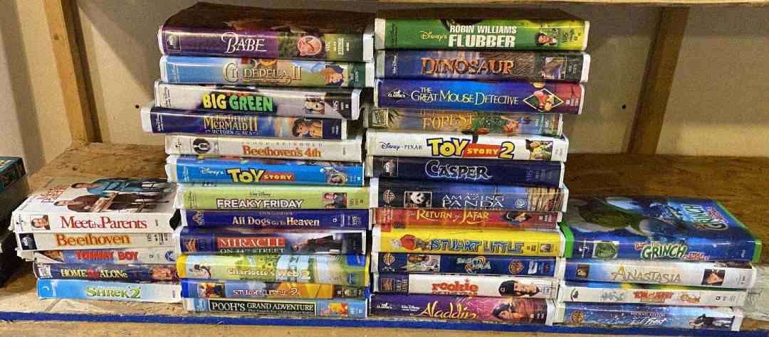 A collection of VHS tapes stacked on wooden shelves, featuring various family movies and animated films such as “Toy Story,” “Flubber,” “The Grinch,” “Shrek 2,” “Aladdin,” and “Casper.”
