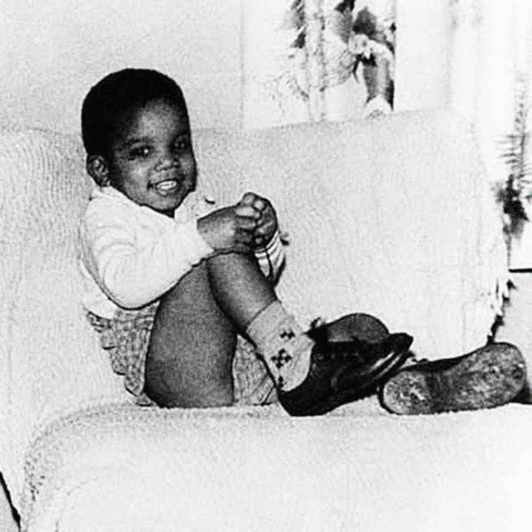A young child sits on a couch, smiling and holding one foot with both hands. The child is wearing a light sweater, patterned socks, and shorts. The background shows a window with curtains. The photo is black and white.