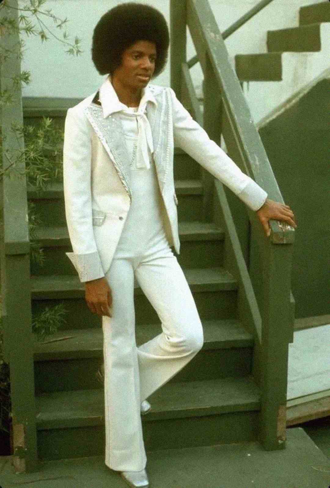 A person with an afro hairstyle stands on outdoor stairs, wearing a stylish white suit with flared pants and a matching shirt, posing with one arm resting on the railing.