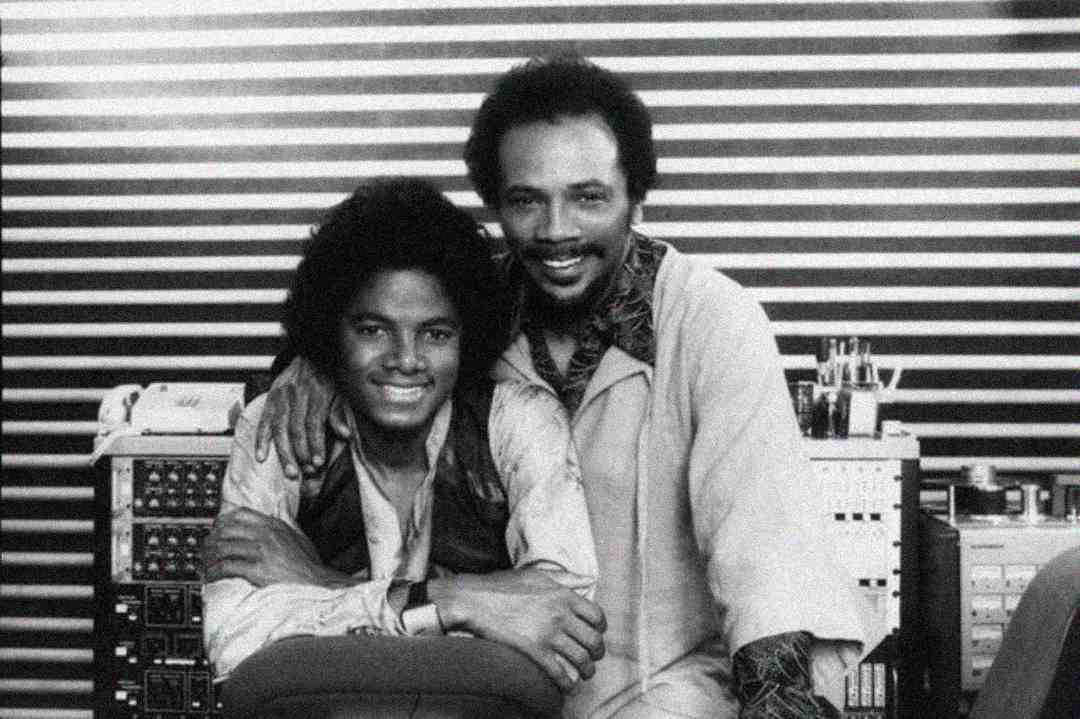 Two men smile and pose together in a recording studio, sitting in front of striped wall panels and audio equipment. One has an arm around the other’s shoulder, both appearing happy and relaxed.
