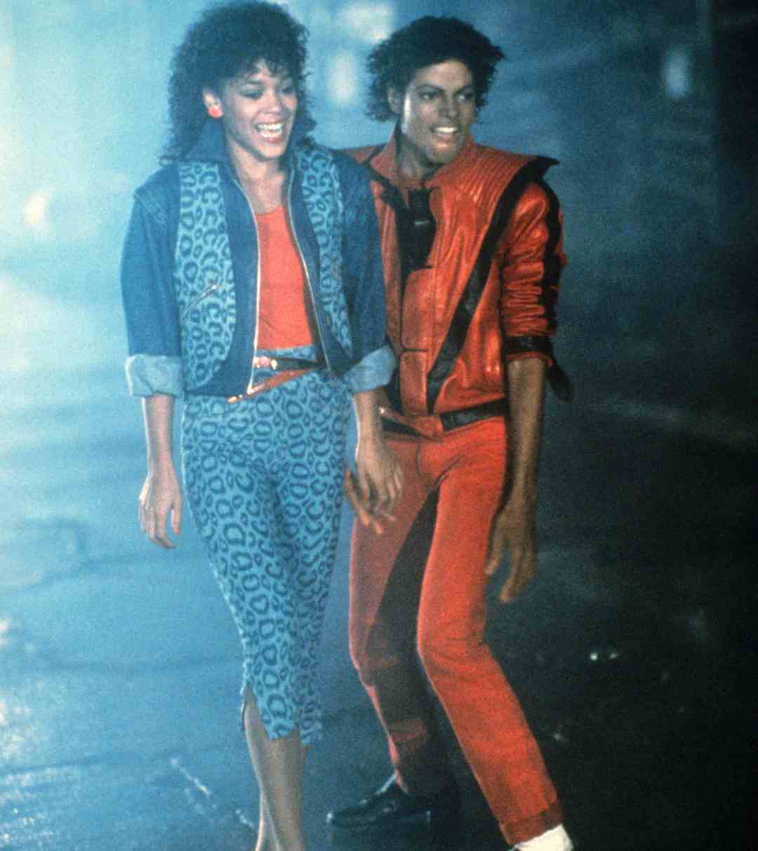 Two people wearing 1980s-style red and blue clothing walk together at night. One has curly hair and a red jacket, while the other wears a blue jacket and leopard-print leggings. They are smiling and holding hands.