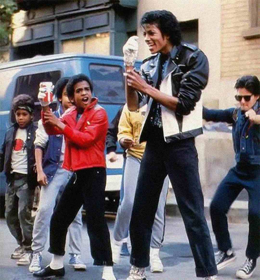 A group of boys dressed in 1980s streetwear dance energetically on a city sidewalk, with the central figure wearing a black leather jacket and performing a dramatic pose with a white glove.