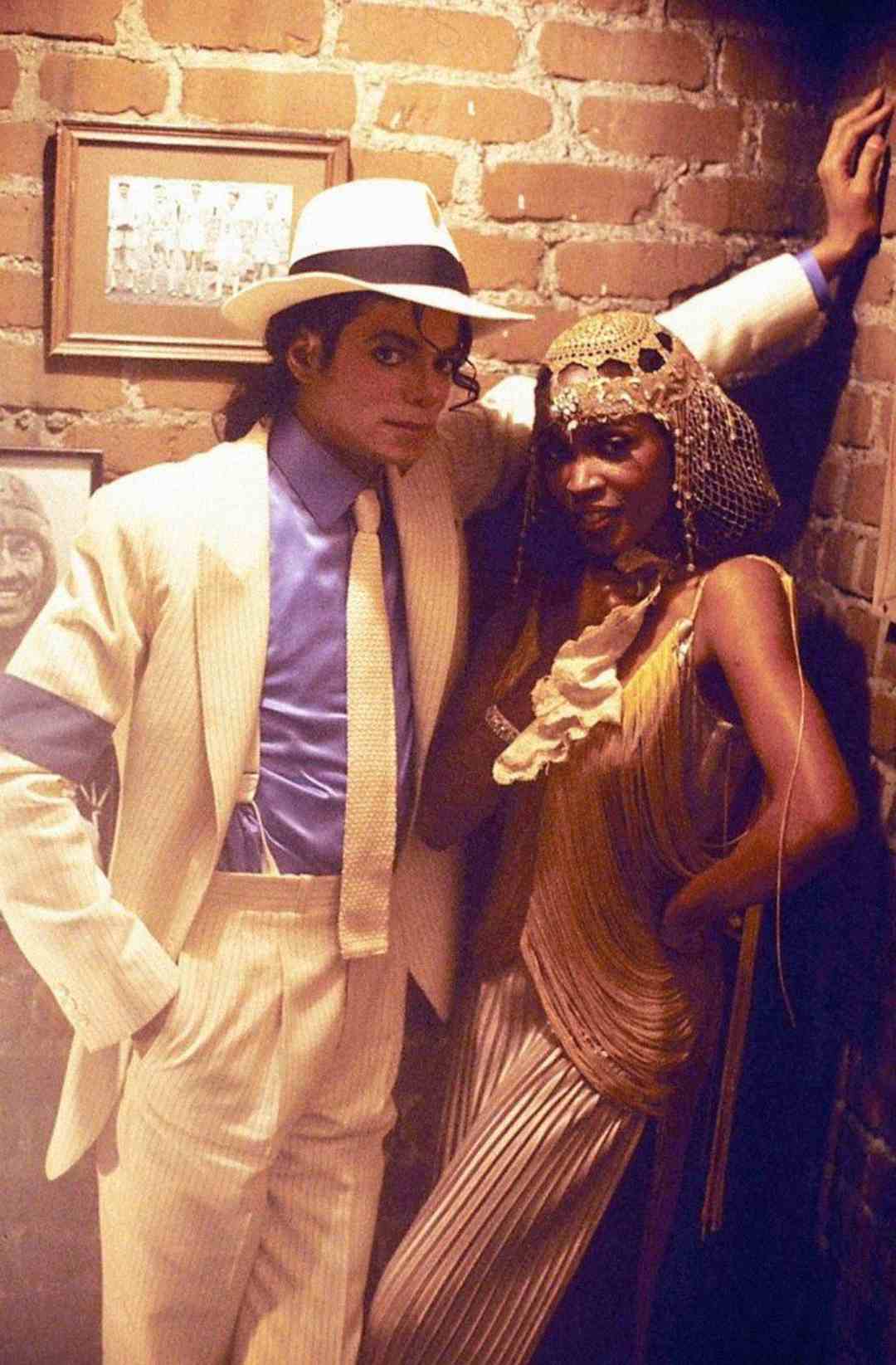 A man in a cream pinstripe suit, fedora, and blue shirt poses beside a woman in a gold flapper-style outfit and beaded headpiece, both standing against a brick wall.