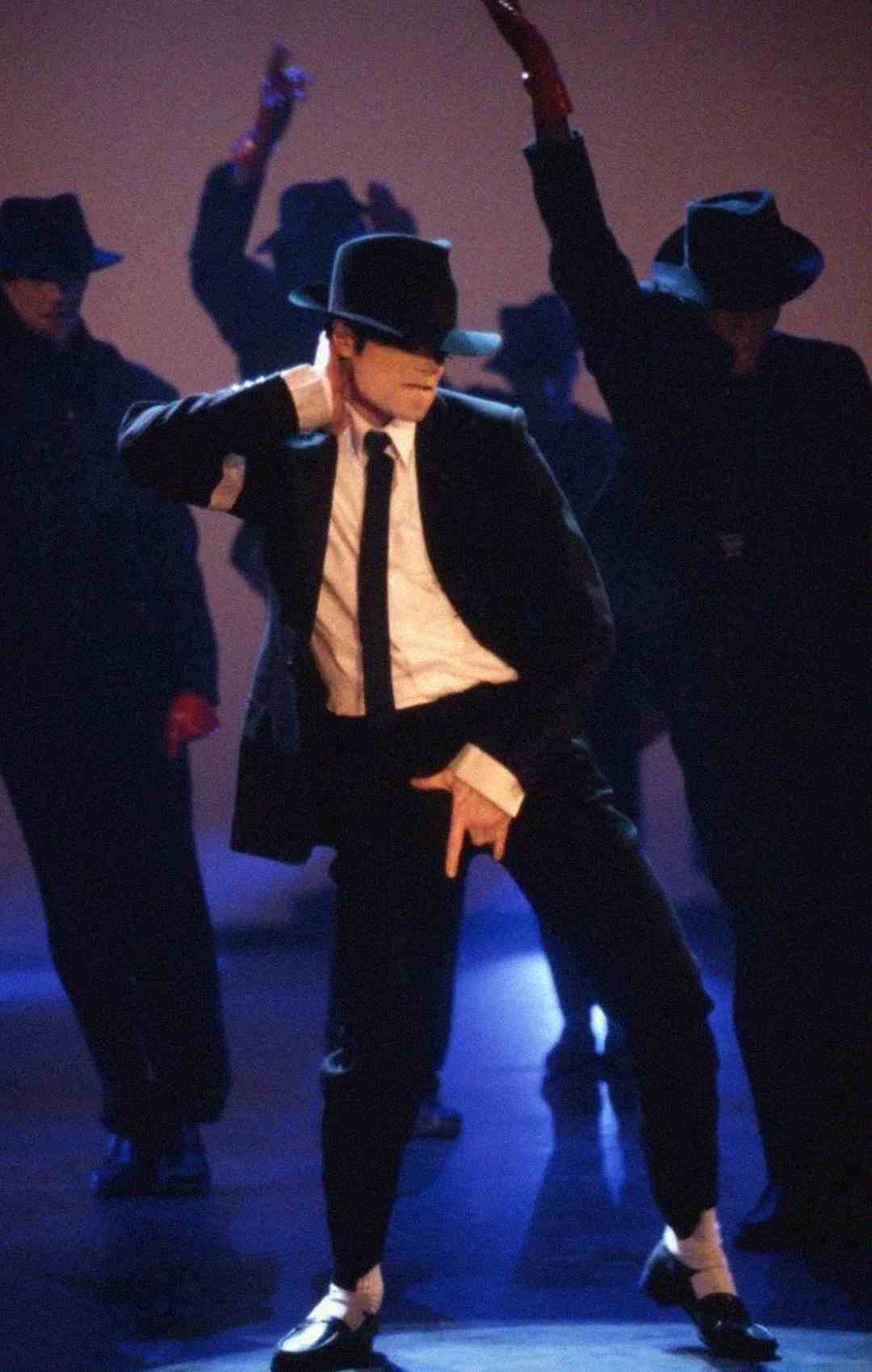 A performer in a black suit, white shirt, black hat, and white socks strikes a dance pose on stage with backup dancers in similar outfits and hats, under dramatic lighting.