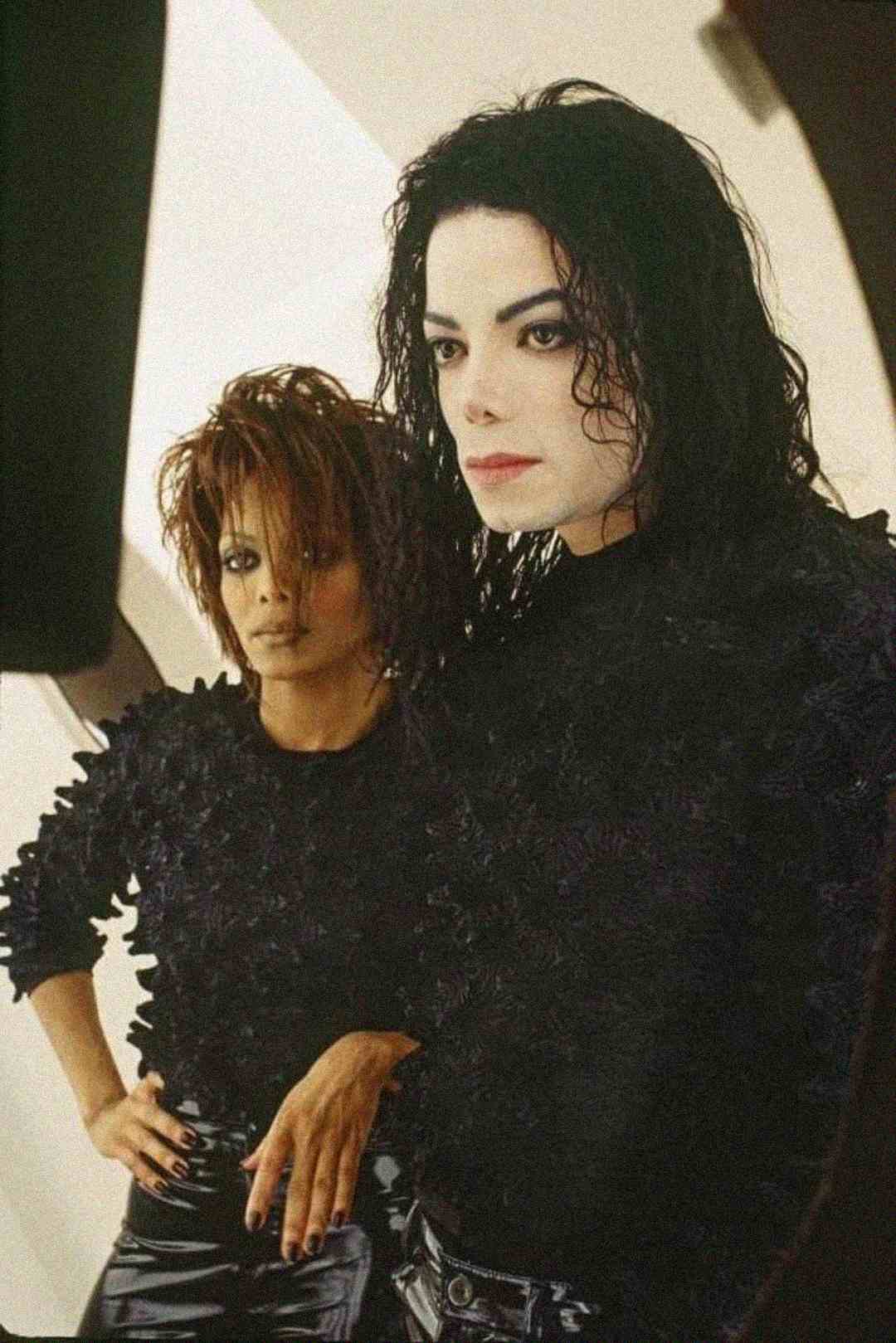 Two people with dark, textured clothing and styled hair stand side by side, looking intently ahead. The person on the right has pale makeup and long, wavy black hair. The person on the left has short, tousled hair and a hand on their hip.