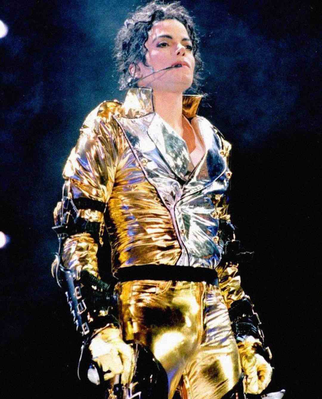 A performer wearing a shiny gold and silver metallic jacket and pants stands on stage with a headset microphone, looking upward under dramatic lighting.