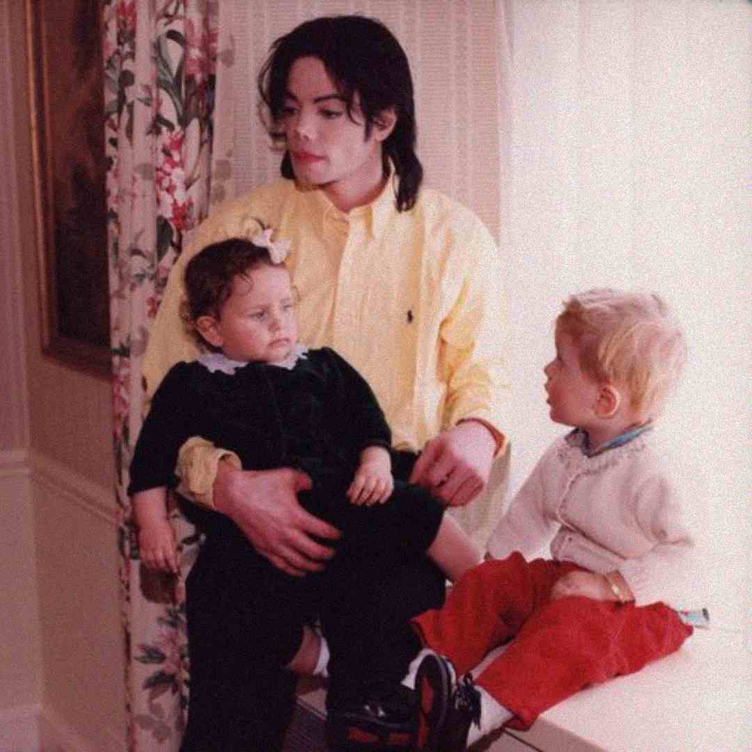 A person with dark hair in a yellow shirt sits indoors by a window, holding a young child in a black outfit while another young child in red pants sits nearby, looking at them.