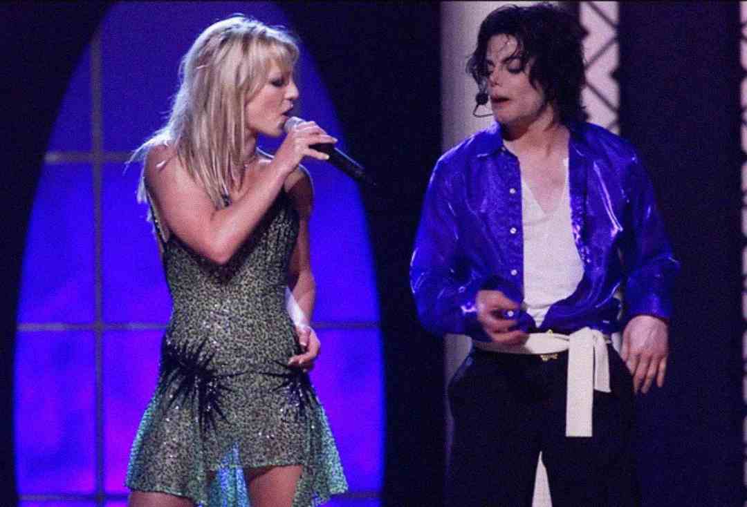 A woman in a sparkly green dress sings into a microphone next to a man in a shiny blue shirt and black pants on stage, both performing under stage lights.