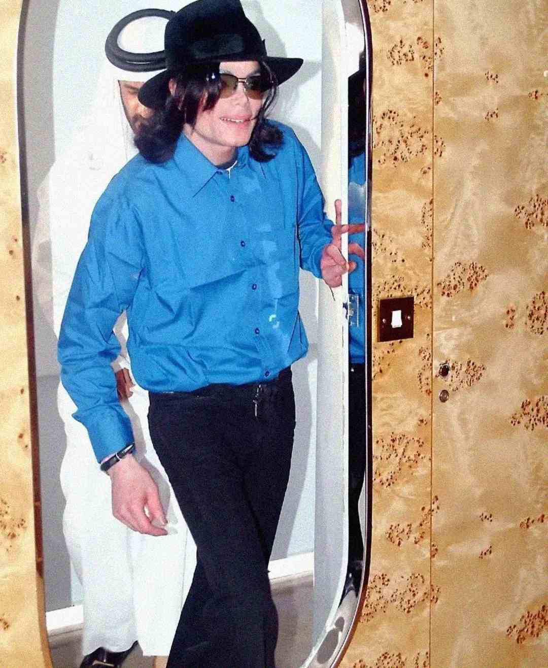 A person wearing a blue shirt, black pants, black hat, and sunglasses is smiling while walking through a doorway. Another person in white traditional Middle Eastern attire stands behind them.