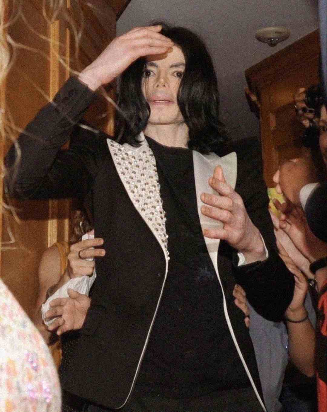 A person with shoulder-length black hair wearing a black jacket with a decorated white lapel is surrounded by people, holding up one hand near their forehead and the other near their chest in a crowded indoor setting.