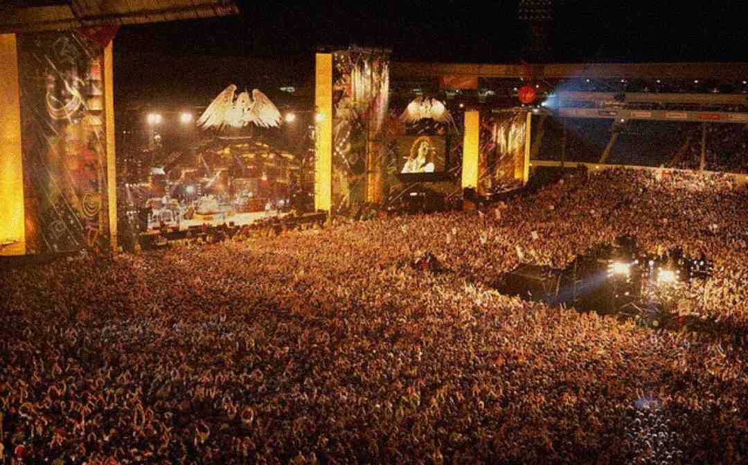 A massive crowd fills an outdoor stadium at night, facing a brightly lit stage decorated with large winged symbols, with performers visible and big screens displaying close-up views.